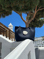 Black handbag with a white flower design on a white railing against a blue sky and tree.