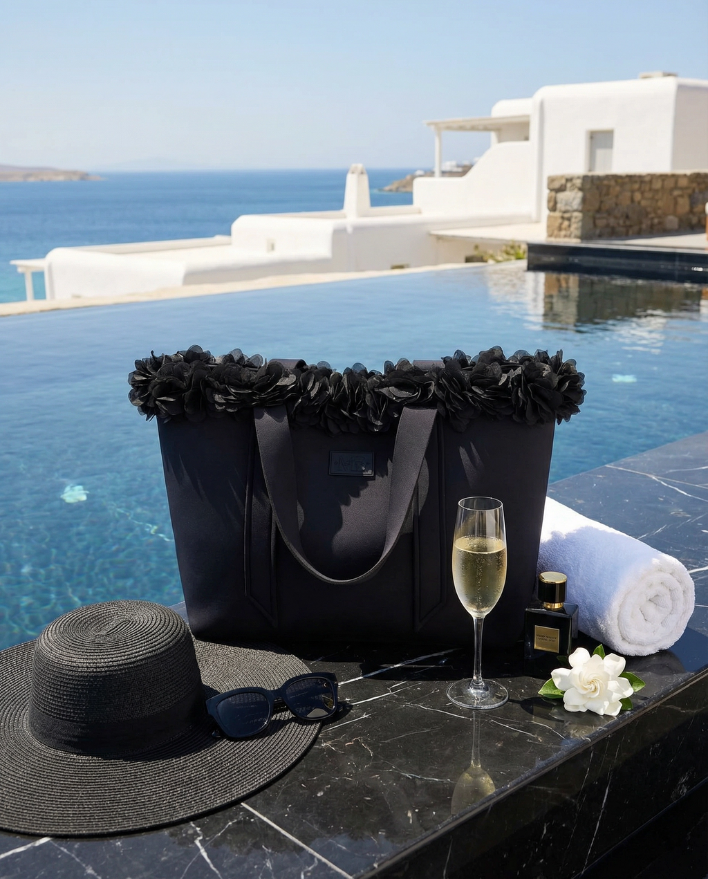 Black tote bag with floral details, black sun hat, sunglasses, glass of champagne, and white towel by a poolside.