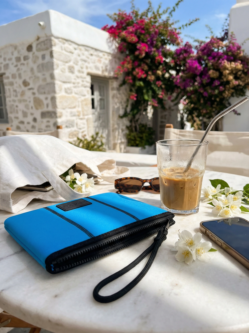 Blue and black pouch on a table with a glass of coffee and phone, outdoor setting with flowers and stone building.