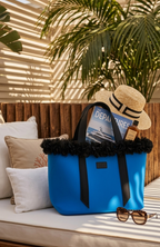 Blue tote bag with black trim, straw hat, and sunglasses on a sunlit outdoor bench.