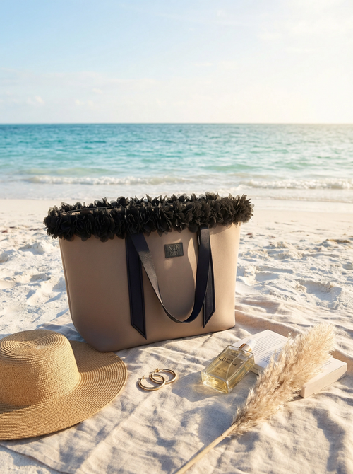 Beige tote bag with black trim, straw hat, and perfume bottle on a sandy beach.