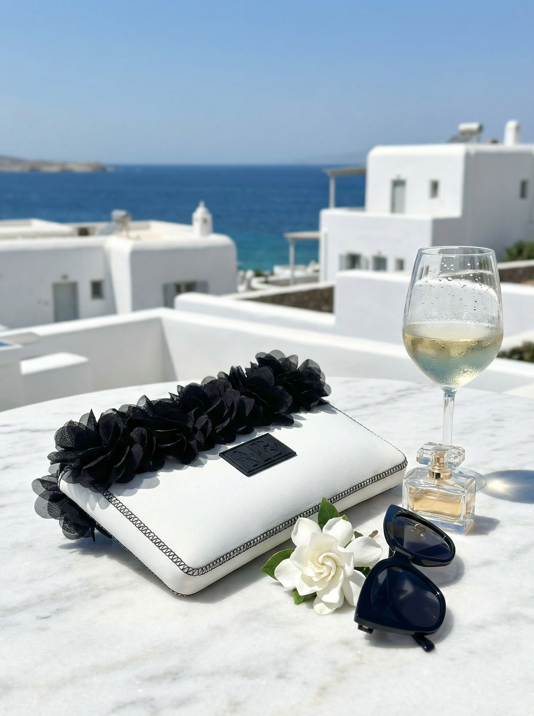 White clutch with black floral details, sunglasses, and a glass of white wine on a marble surface with ocean view.
