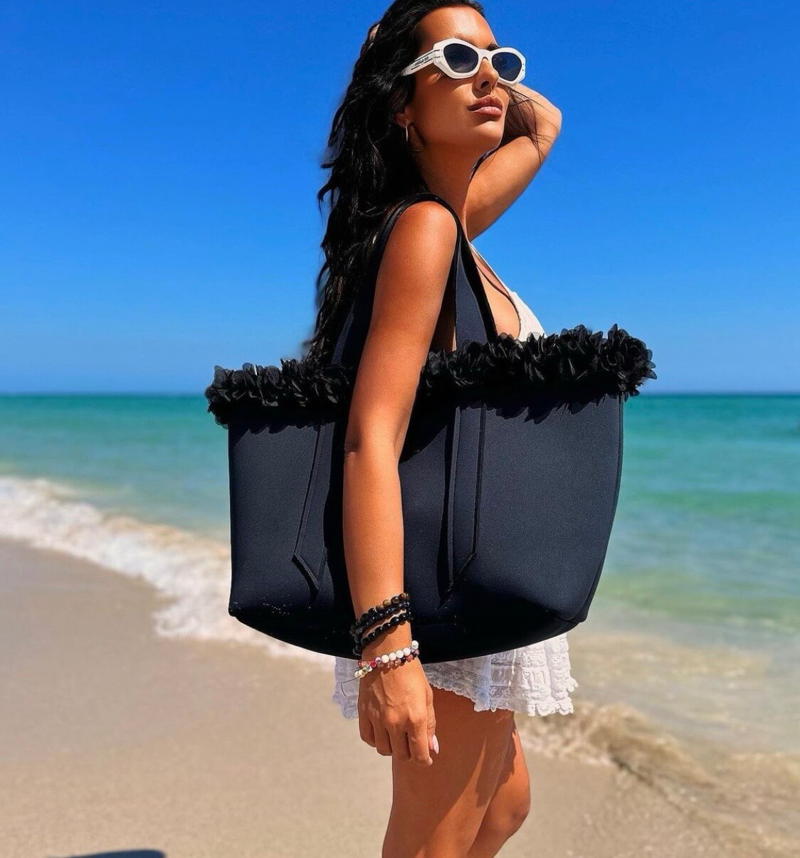 Woman on a beach holding a large black tote bag with ruffles, wearing sunglasses.