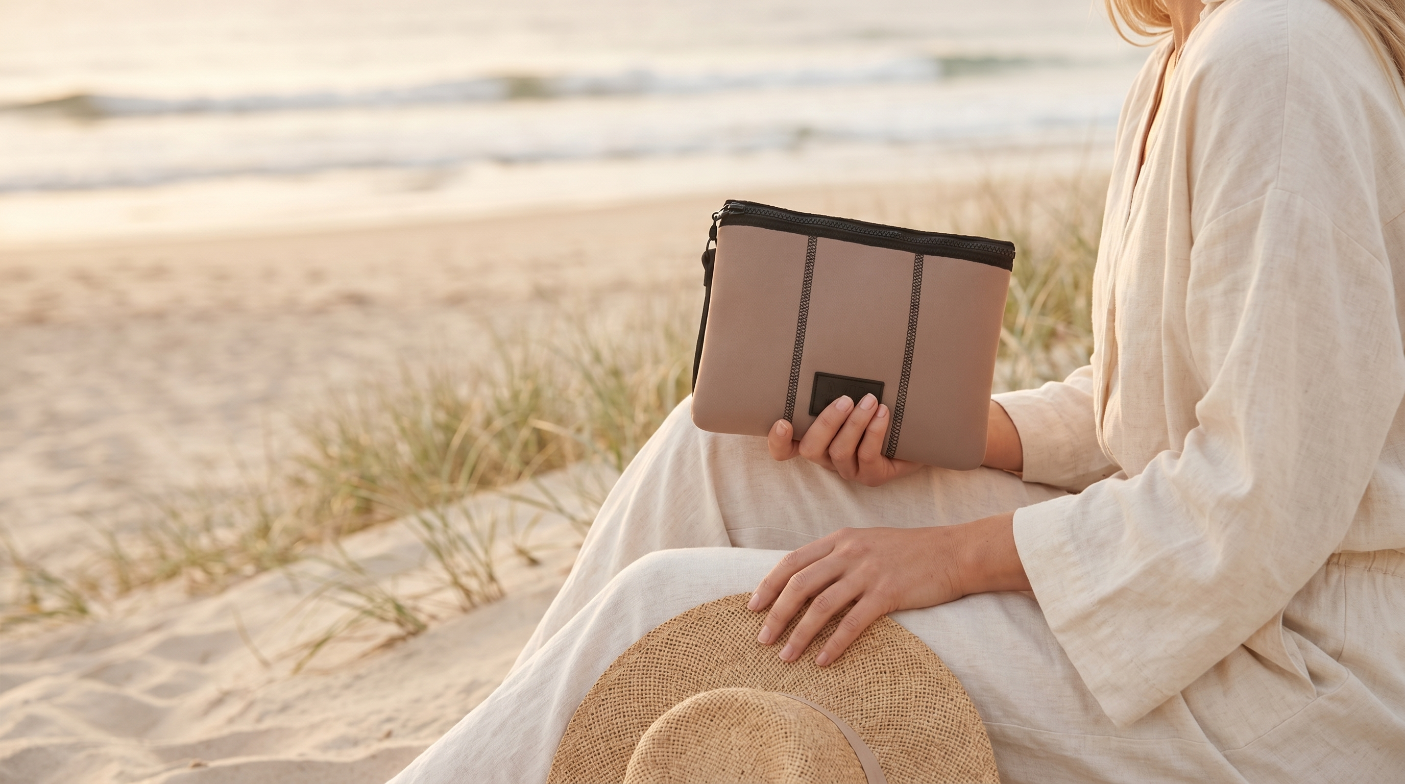 Person sitting on a beach holding a beige clutch bag with a straw hat beside them.