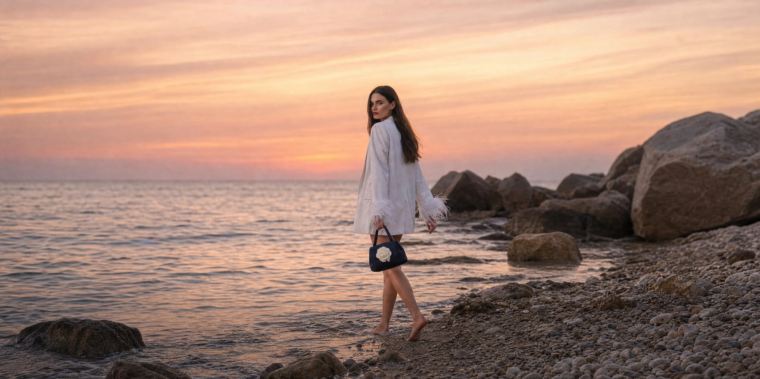 Woman in a white dress standing on a rocky beach at sunset