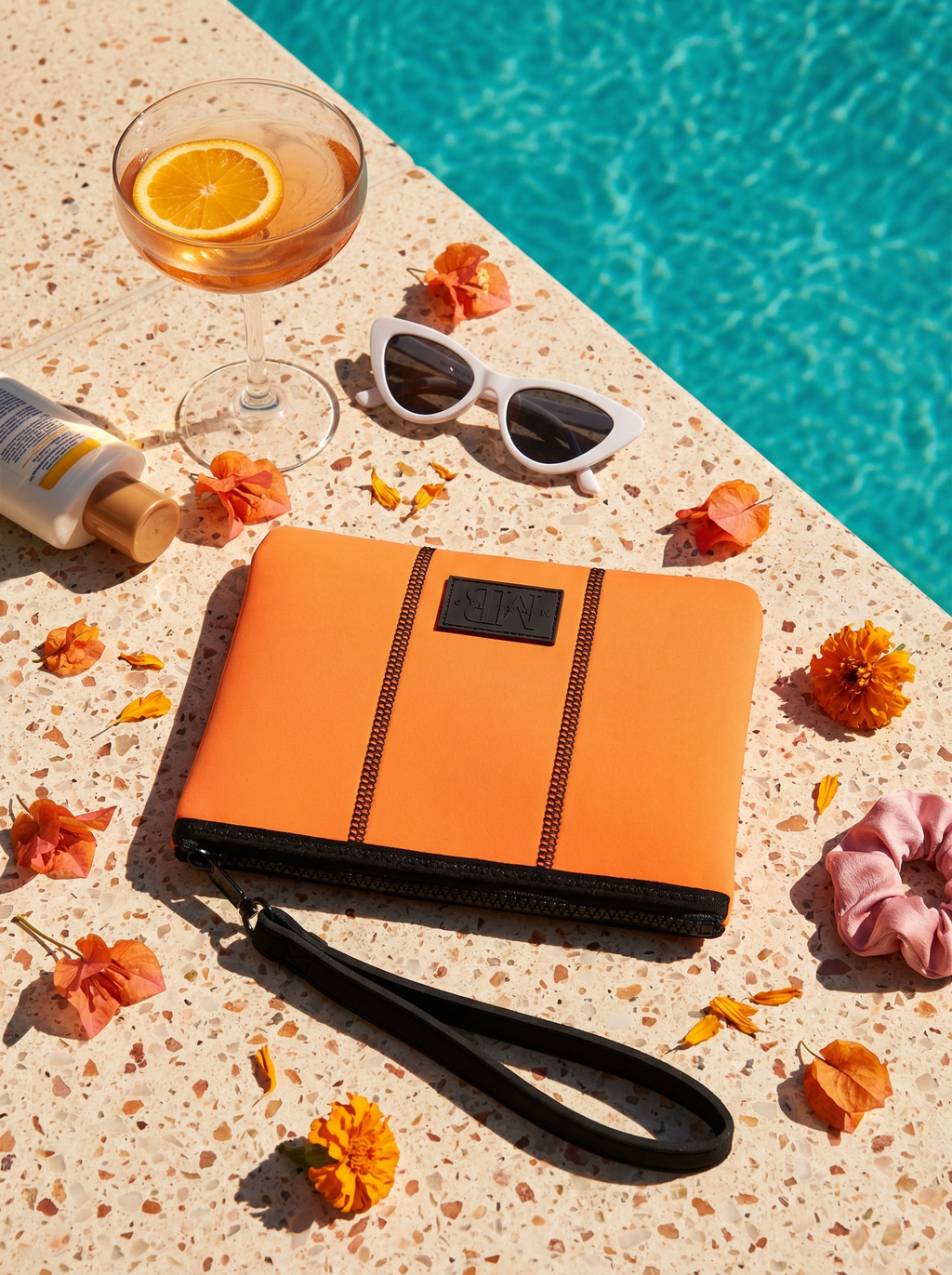 Orange clutch bag with black strap on a poolside surface with flowers and a drink.