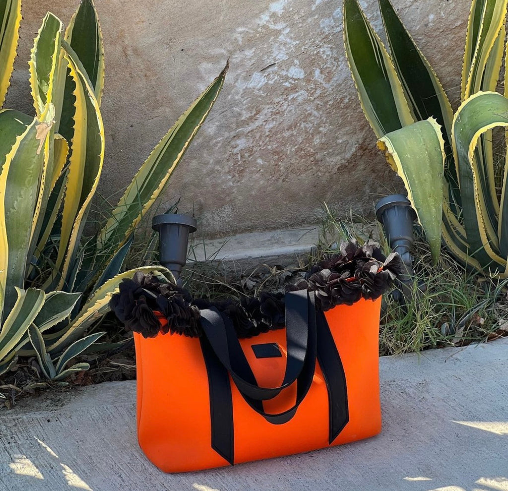 Orange tote bag with black handles and trim on a stone surface with plants in the background