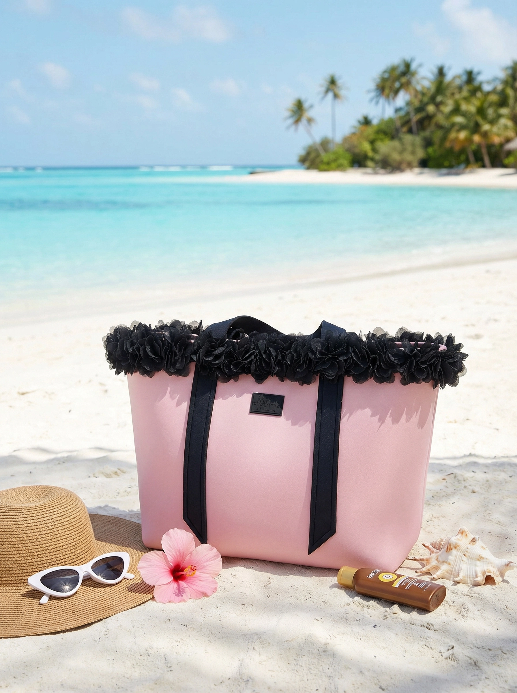 Pink beach bag with black floral details on a sandy beach with a hat, sunglasses, and flower.