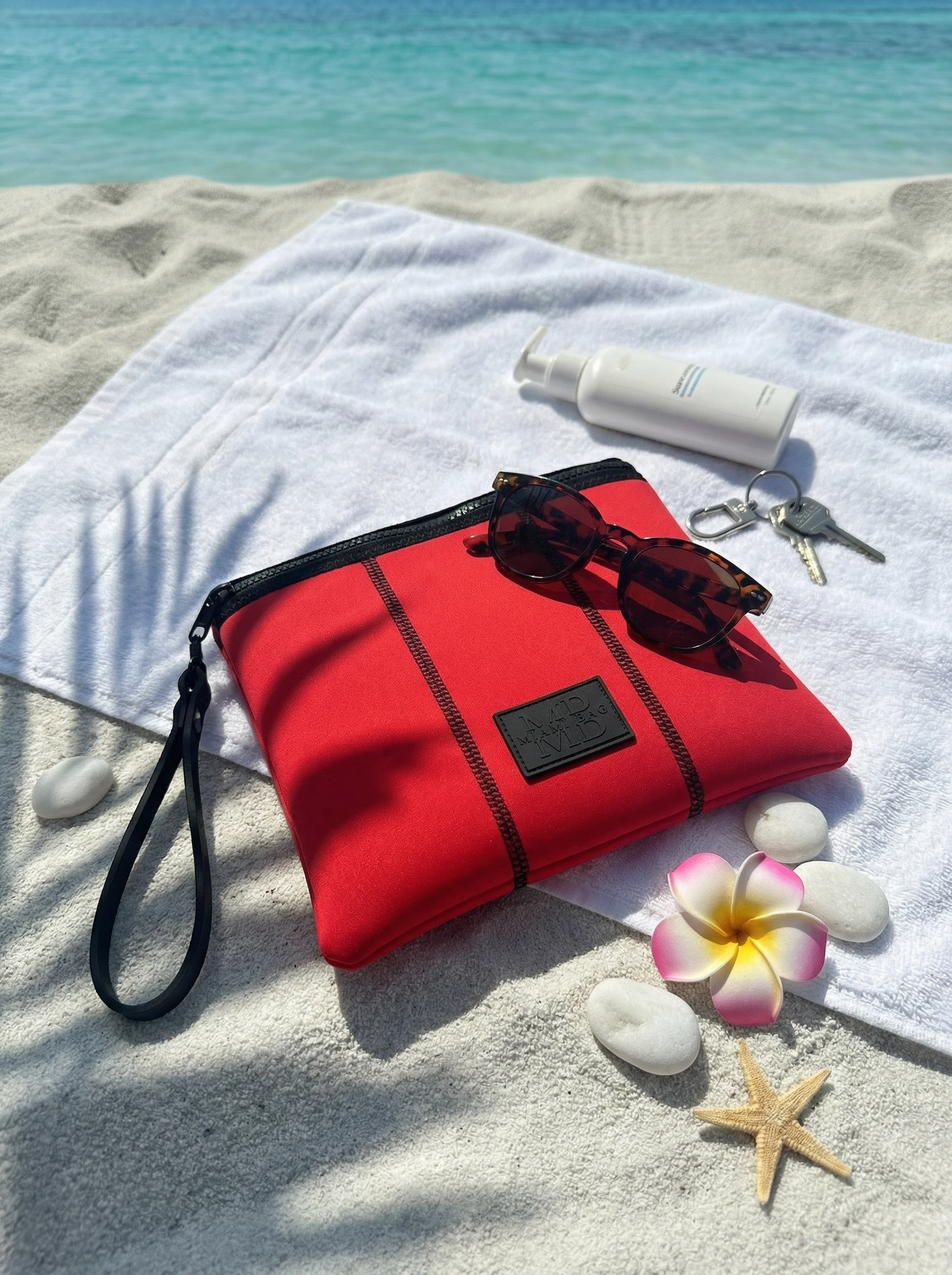 Red bag, sunscreen bottle, keys, sunglasses on a towel with beach items.