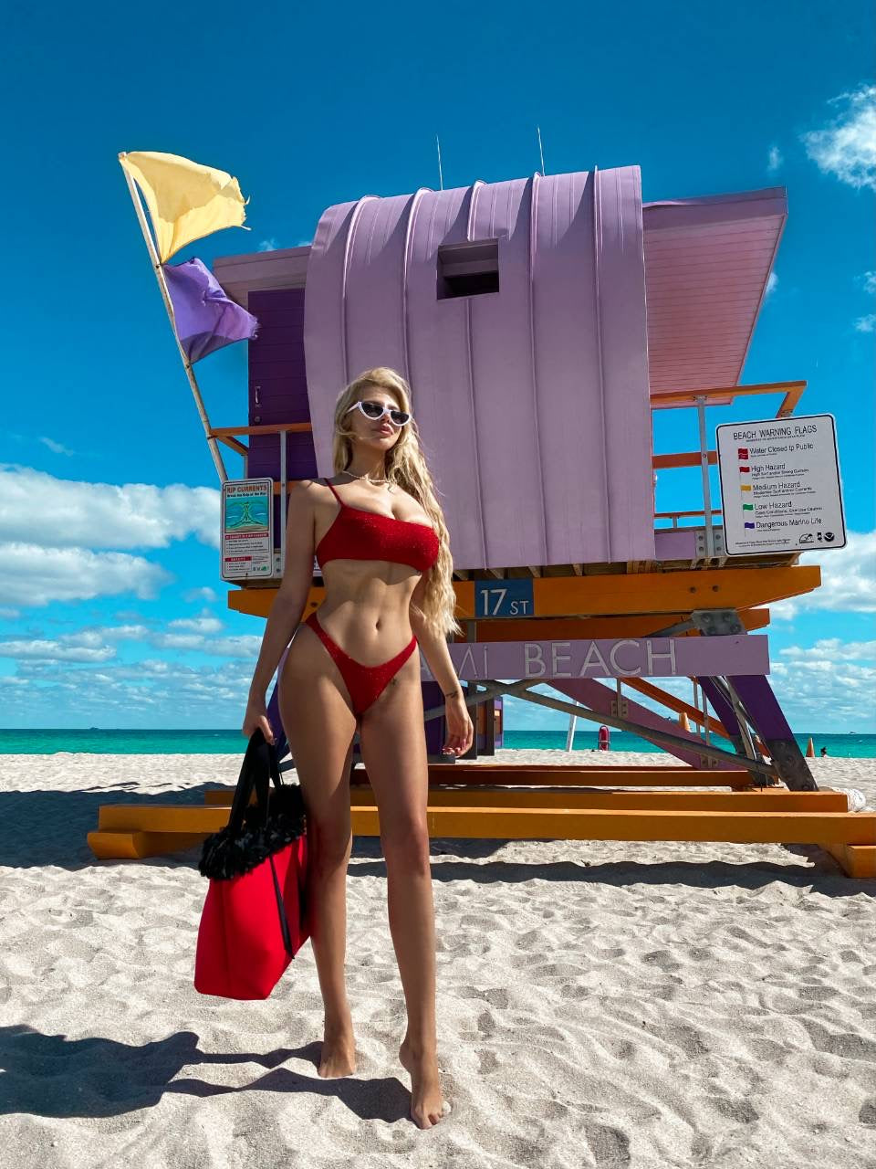 Woman in a red bikini standing on a beach with a colorful lifeguard stand in the background.