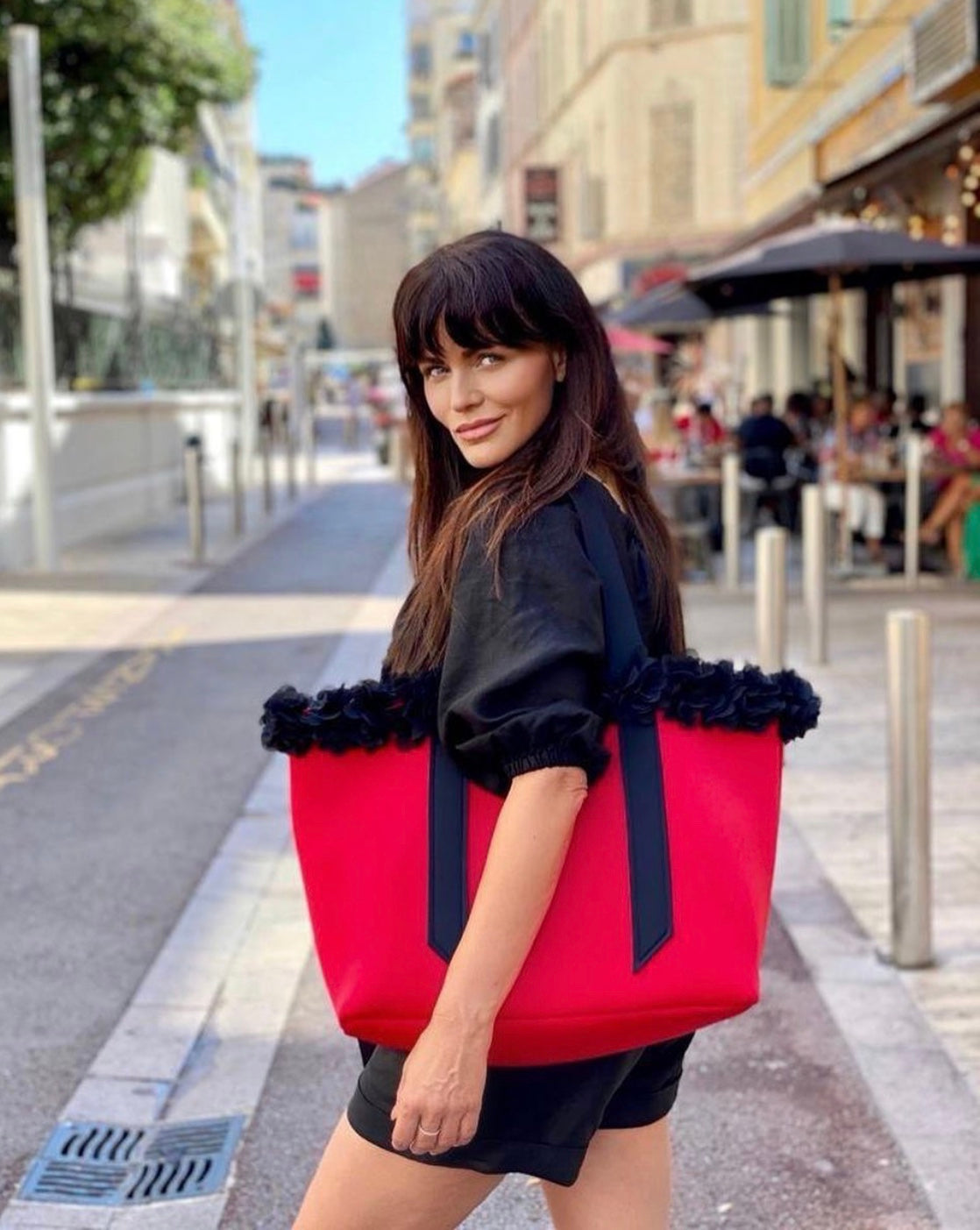 Woman holding a red tote bag with black trim on a city street.