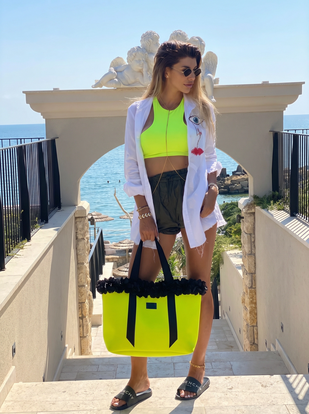 Woman in bright yellow top and shorts with a matching bag, standing on steps with ocean view.
