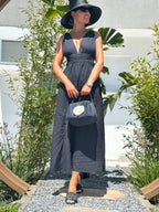 Woman in a long dark dress and hat standing outdoors with plants, holding a black bag.