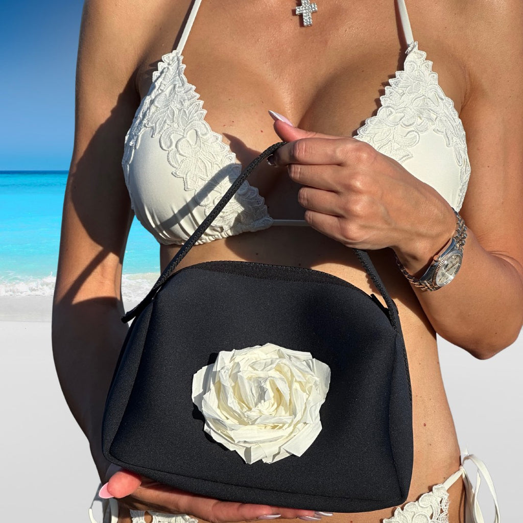 Person holding a black handbag with a white rose design on a beach.